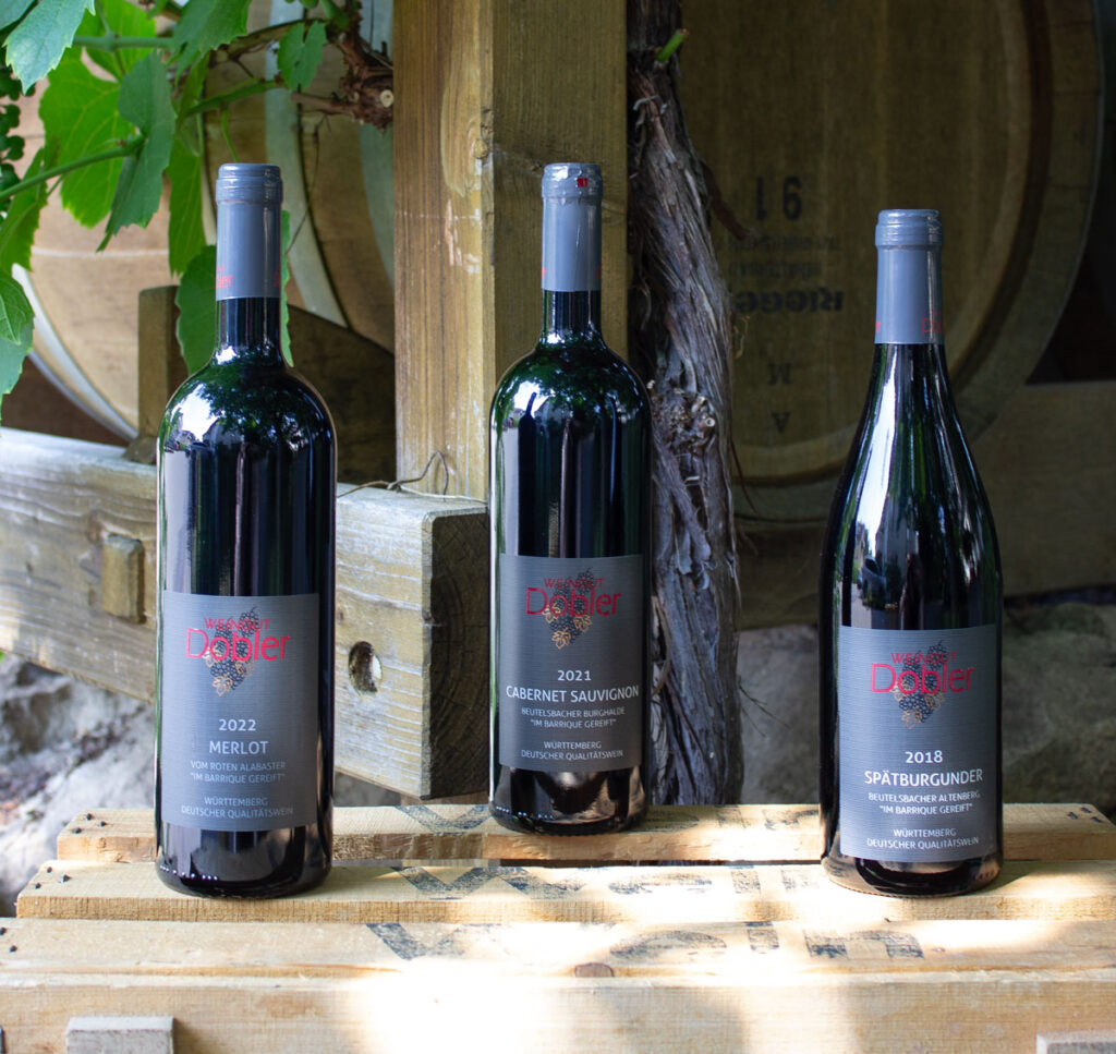 Wein-Probepaket aus dem Barriquefass mit allen drei Weinflaschen aus dem Weinpaket auf einer Holzkiste vor Barrique-Fässern mit Weinranken und Steinen.