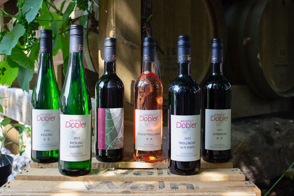 Wein-Probepaket Remstal Pur mit allen sechs Weinflaschen auf einer Holzkiste. Im Hintergrund sind Barriquefässer, Weinranken und Steine zu sehen.